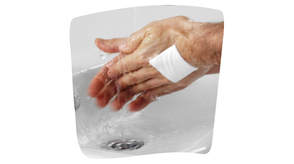 Imagen de una persona que se está lavando las manos en el lavabo del baño con un apósito impermeable que cubre una herida sobre el dorso de la mano izquierda. Imagen de una persona que se está lavando las manos en el lavabo del baño con un apósito impermeable que cubre una herida sobre el dorso de la mano izquierda.